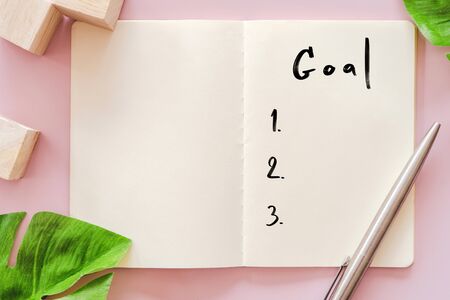 Goal list on notebook with pen and green leaves. Pastel color background.