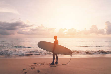 Man hold surfboard standing at tropical sunset beach background. Summer vacation and sport adventrue...