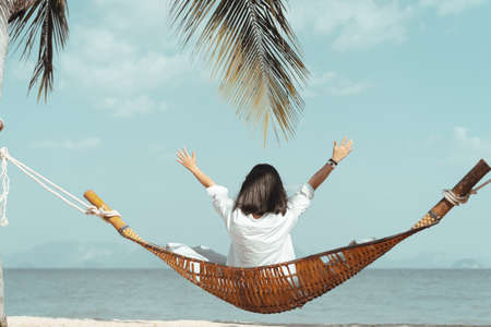 Happy woman hand raising and sitting on hammock at tropical beach background. Summer vacation and freedom feel good concept. Vintage tone color style.の写真素材