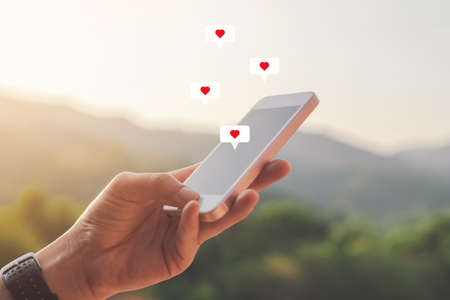 Close up woman hand hold using smart phone with heart icon at top of mountain and sunset sky...