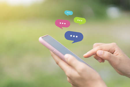 Close up woman hand hold using smart phone with chat icon flying at outdoor nature park background. Copy space of technology business and travel holiday concept. Vintage tone filter effect color.の写真素材
