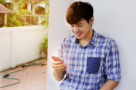 Young thai man holding mobile phone, Asian man using smartphone, Cheerful asian model next to home...