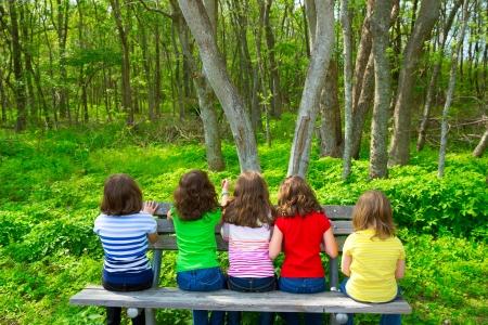 Children sister and friend girls sitting on park bench looking at forest rear viewの写真素材