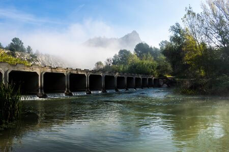 Moody landscape of a river crossed by a bridge with the mountains at the foregroundの写真素材