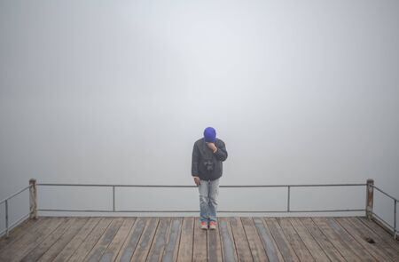 Young man standing alone on wooden edge of footbridge at lake. Mist over water. Foggy air. Early...