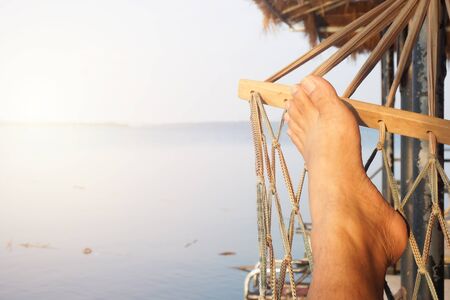 relaxing time concept,summer concept,Man relaxing on over water hammock in water villa bungalow. Feet view overlooking tropical sea from Island.の写真素材