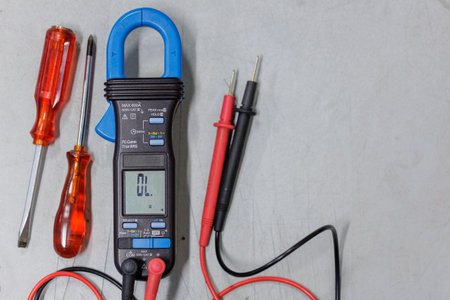 A blue and black digital clamp meter is placed on a light-colored workbench next to two red-handled screwdrivers. This image represents tools of the electrical trade, symbolizing diagnostics, repair, and professional maintenance.の写真素材