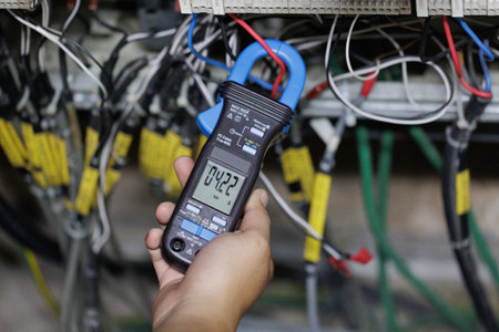 A professional's hand holds a digital clamp meter, with the probes connected to a circuit, showing a voltage reading of 04.22. The image depicts the essential task of a technician performing electrical checks in a complex wiring cabinet.の写真素材
