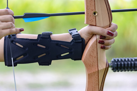 Detailed shot of a woman's hand with red nail polish firmly gripping a wooden recurve bow, wearing a protective arm guard, captured in the ready-to-shoot position.の写真素材