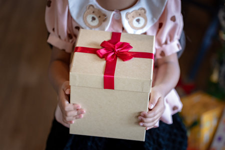 A focused, close-up shot of a child's hands holding a simple but large square cardboard gift box, tightly secured with a vibrant red satin bow. Represents the moment of giving a cherished holiday presentの写真素材