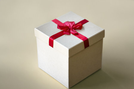 A clean, square beige gift box tied with a vibrant red ribbon bow, placed on a warm cream-colored...
