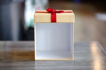 A square, empty beige gift box standing open on a gray surface with the lid (featuring a small red...
