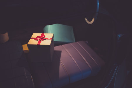 Focus on a beige gift box with a red bow sitting on a purple suitcase inside the dark car trunk, suggesting holiday travel preparations and the effort of delivering presents to loved onesの写真素材