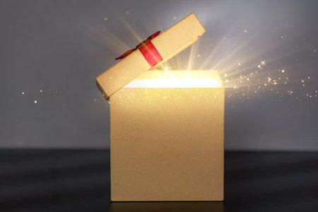 A single beige gift box standing on a dark surface with the lid raised, revealing a powerful burst of shimmering light and glitter. Perfect visual for surprise, prize, or a special Christmas present revealの写真素材