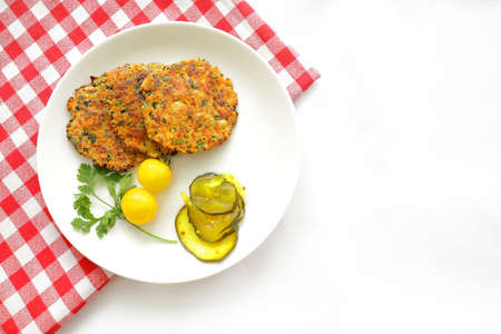 Zucchini patties on white plate garnished with tomatoes, cilantro and pickles against a red checkered cloth.の写真素材