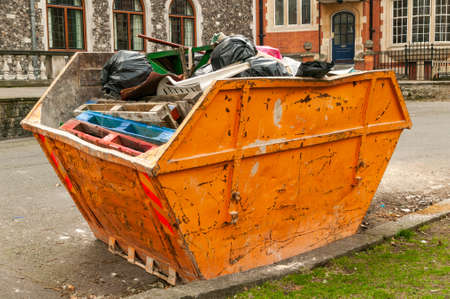 Heavy metal orange iron skip container full of scrap waste household rubbish such as black plastic bin bags and pallets all destined for a landfill site, which causes environmental pollutionの写真素材