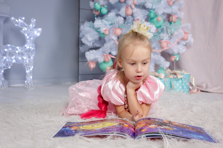 Portrait of cute european little blonde princess girl with crown in beautiful dress lying on the floor and reading book in decorated studio in christmas location with toys and christmas treeの写真素材