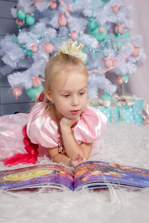Portrait of cute european little blonde princess girl with crown in beautiful dress lying on the floor and reading book in decorated studio in christmas location with toys and christmas treeの写真素材