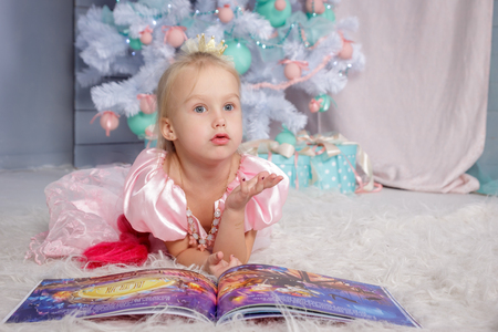 Portrait of cute european little blonde princess girl with crown in beautiful dress lying on the floor and reading book in decorated studio in christmas location with toys and christmas treeの写真素材