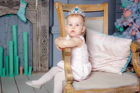 Portrait of a cute little european blond princess girl with a crown  in a beautiful dress sitting on a vintage chair in a studio decorated in the Christmas theme with toys, christmas tree and fireplaceの写真素材