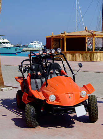 An orange beach buggy parked at a marina with luxury yachts in the background