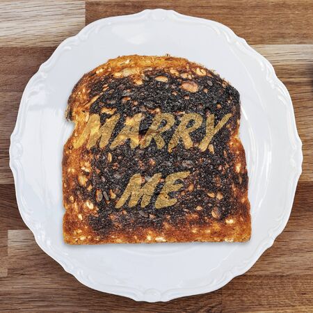 A slice of burnt toast with a marraige proposal message on a shiny white plate placed upon a wooden...