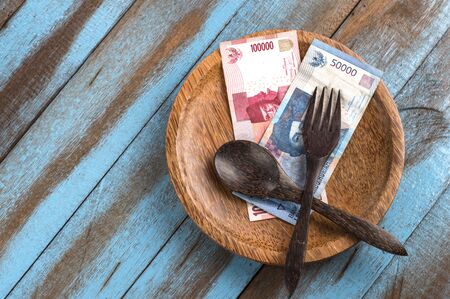 brown wooden spoon and fork as well as the Indonesian currency banknotes on the wooden plate,...