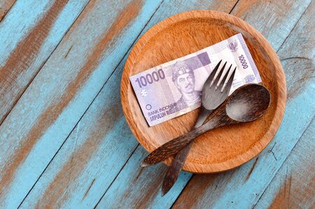 brown wooden spoon and fork as well as the Indonesian currency banknotes on the wooden plate, illustrating expensive or cheapening the cost of eatingの写真素材