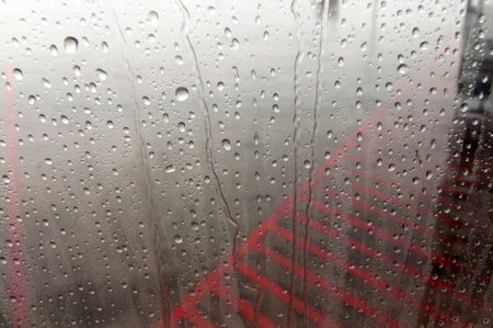 Raindrops cling to glass, obscuring a red structure. A gloomy day outside creates a moody, abstract...