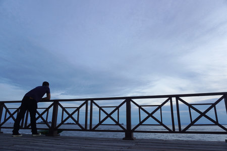 A solitary figure gazes at the expansive ocean from a wooden pier as dusk settles, evoking feelings of peace and introspection. Perfect for travel and lifestyle themes.の写真素材