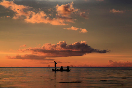 Silhouetted figures sail a tranquil sea during a breathtaking sunset, painting the sky with vibrant oranges and golds.の写真素材