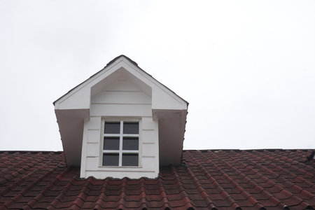 A sharp, modern dormer window with a clean white frame stands out against the textured red tiles of a classic pitched roof, set against a bright, overcast sky.の写真素材