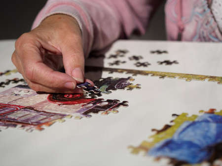 Close view of a female hand with jigsaw piece about to be placed into a puzzle on white table.Lockdown activity.Imageの写真素材