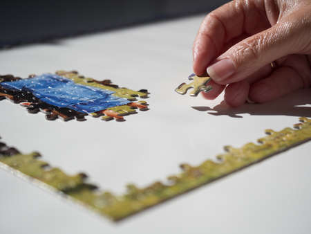 Close view of a female hand with jigsaw piece about to be placed into an incomplete puzzle on white table.Lockdown activity.Imageの写真素材