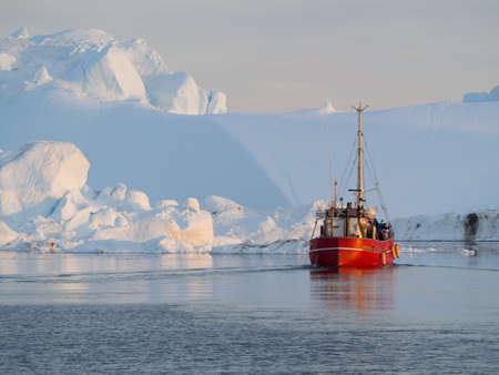A red fishing boat motors through tranquil sea surrounded by icebergs.Greenland.Arcticの写真素材