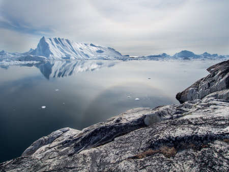Iceless rock in the foreground contrasting with huge glacial ice forms in the distance across a tranquil ocean bay in Greenland highlights climate change.Imageの写真素材