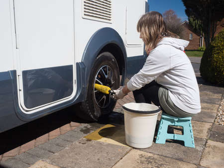 A lady motorhome owner cleans the wheels with a yellow wheel brush.She has a bucket beside her as she sits on a stoolの写真素材