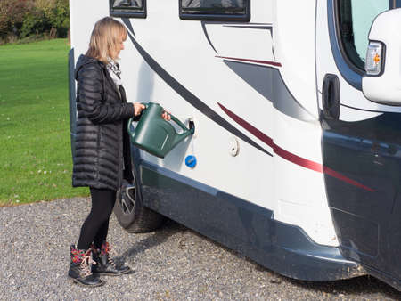 A lady stands refilling the drinking water tank of her motorhome using a green watering can.The blue filler cap hangs down as the fresh water is poured into the side of the vehicle.の写真素材