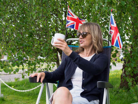 A lady sits in a camping chair in her garden and gestures cheers with a mug in her hand.Two union jack flags are behind her in the garden with tree in background.の写真素材