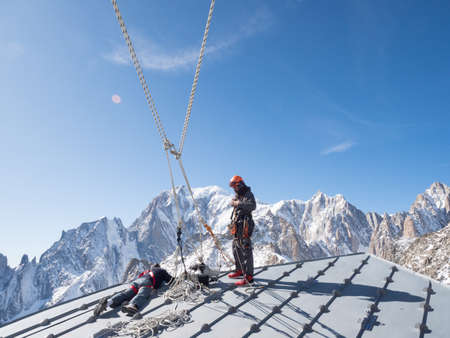 A climber in full safety gear prepares to abseil from a rooftop to make repairs to the cable car station with Mont Blanc in the background.Safety lines appear dramatically from top of imageのeditorial素材