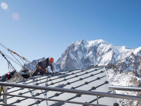 A climber in full safety gear climbs over the edge of a rooftop to make repairs with dramatic backdrop of Mont Blanc in the backgroundの写真素材