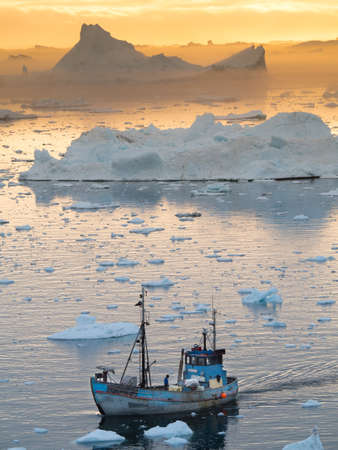 A Greenland fishing boat moves through melting sea ice with drifting icebergs in background lit by orange glow of sunset.Climate Crisis.Vertical Imageの写真素材