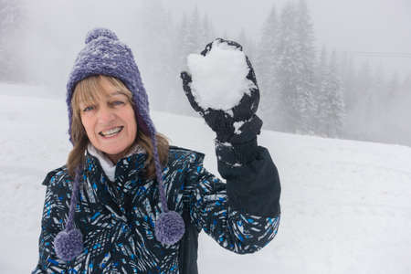 A lady holds a snowball in a gloved hand.She is laughing and looks about to throw it towards camera.She wears a purple woollen hat with tassels and baubles and a ski jacket.Snow and trees.の写真素材