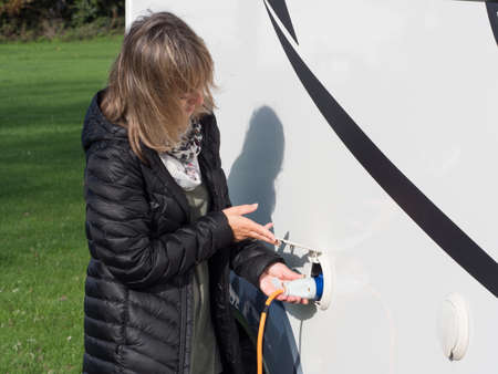 A lady lifts a cover flap and plugs in an orange electric hook up lead into the side of her motorhome to provide power to the camper van.Recreational Vehicleの写真素材