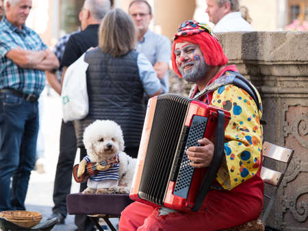 A smiling busker dressed as a colourful clown plays an accordion.The street performer has a dog by his side which has been dressed to look as though it is playing a guitar.Both are seatedのeditorial素材
