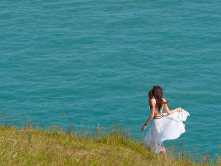 A young woman swirls a flowing white dress as she walks on a grassy sea cliff with aqua sea in the background.Imageのeditorial素材