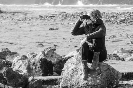 An attractive female sits on a rock looking through binoculars.Coast and sea in background. Monochromeの写真素材