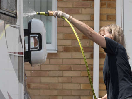 A lady motorhome owner stands cleaning her recreational vehicle with a hose pipe.Spray can be seen from the pipeの写真素材