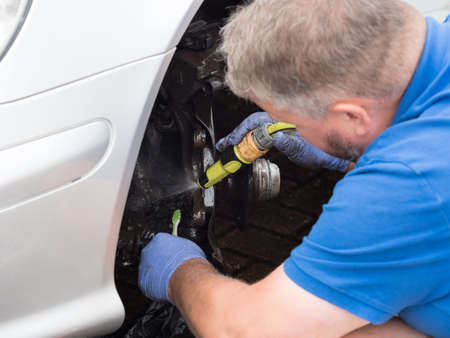 A male mechanic cleans the wheel mount of a car with a hose pipe spray.He wears blue gloves and uses a toothbrush.Close viewの写真素材