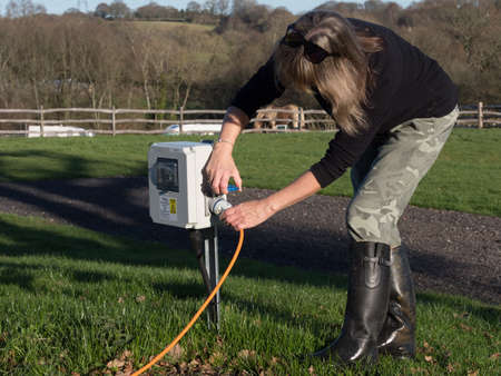 A female motorhomer plugs the orange electric hook up lead into a campsite supply point using the blue connector.She wears black rubber boots and camouflage trousers on sunny day with sunglassesの写真素材
